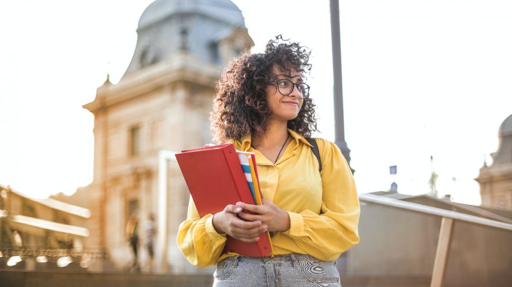 Une jeune fille est dehors, devant un bâtiment. Elle regarde au loin, l'air confiant. Elle tient des classeurs et cahiers dans ses bras.