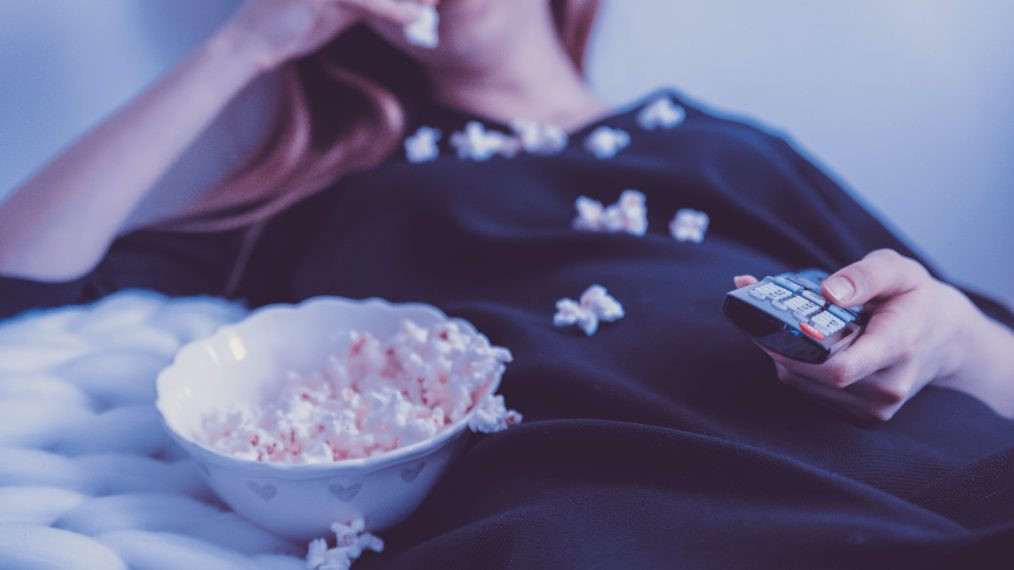 Une femme est allongée et tient des popcorn et une télécommande. Elle semble binge watcher une série !