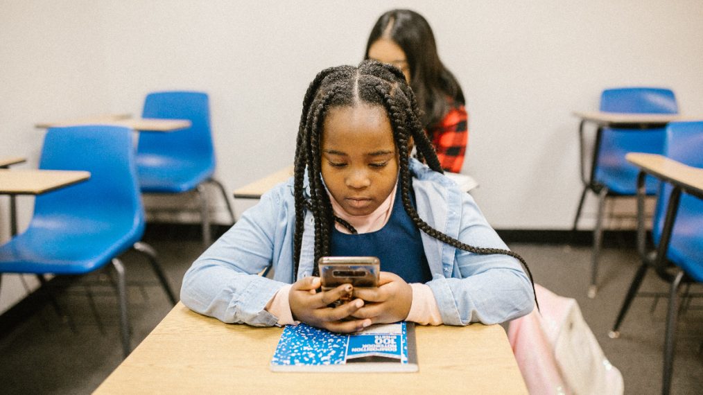 Une petite fille est dans une salle de classe, assise à son bureau. Elle a les yeux rivés sur son téléphone et semble triste.