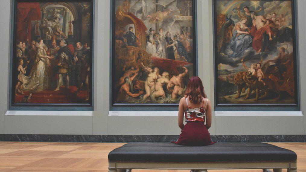 Une jeune fille est assise de dos dans un musée. Elle est face à trois grands tableaux.